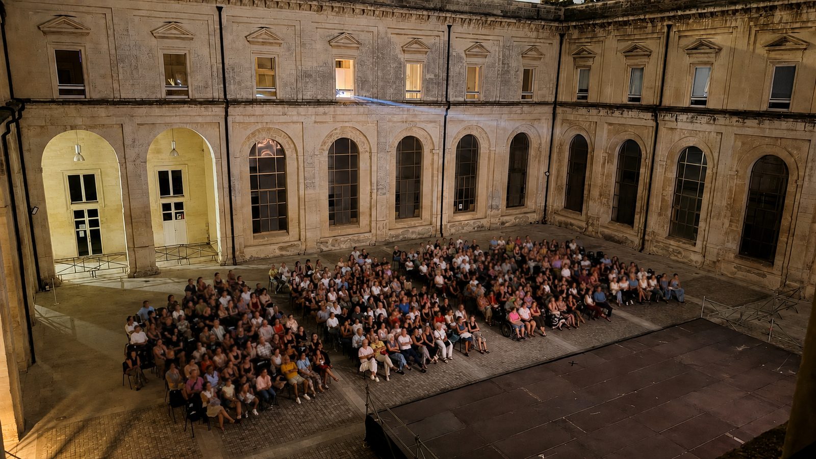 Il chiostro di sera con la platea gremita davanti al palco, le arcate e le finestre illuminate sullo sfondo