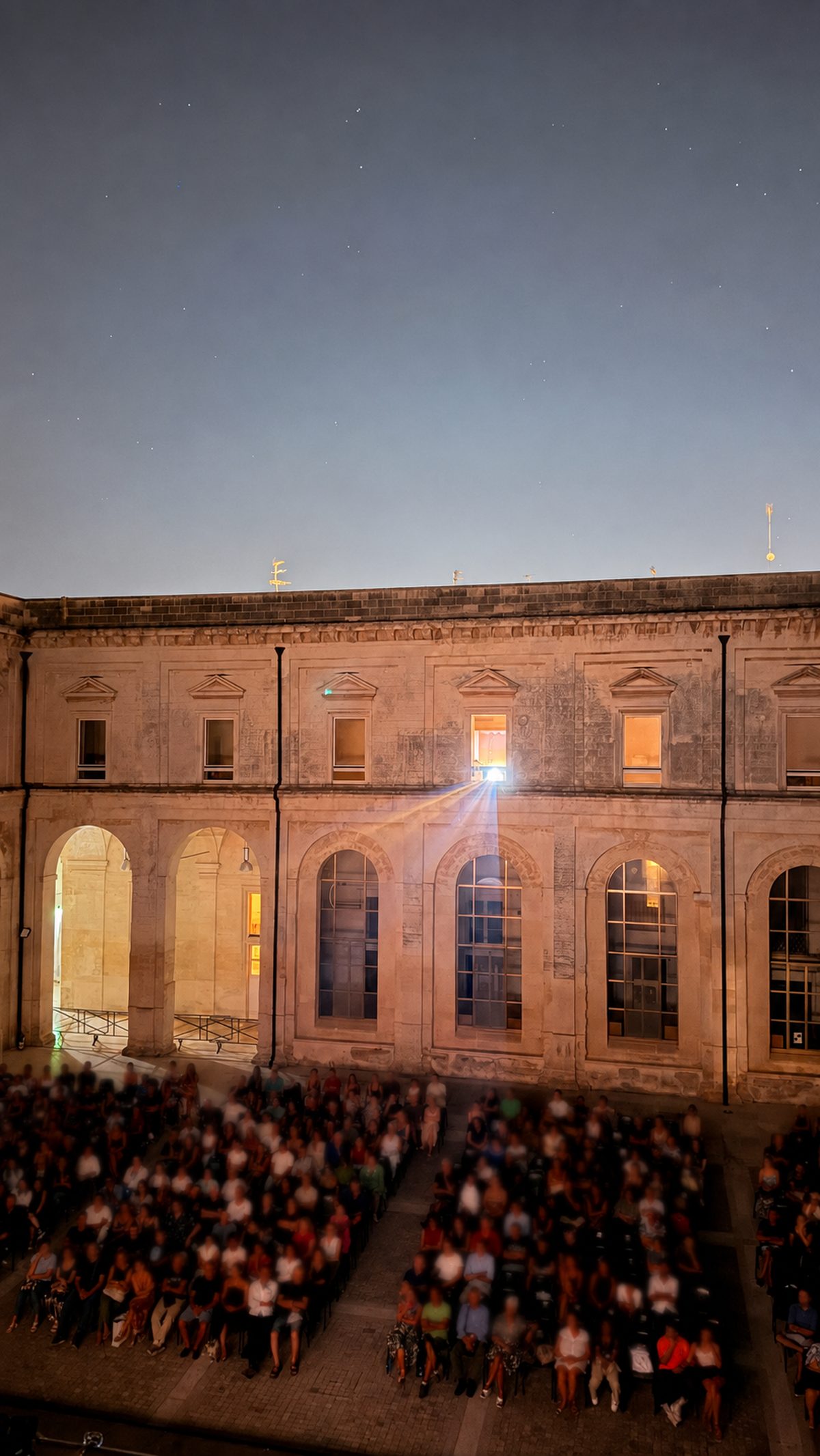 Il fascio di luce del proiettore che esce da una finestra del primo piano e attraversa il chiostro sopra il pubblico