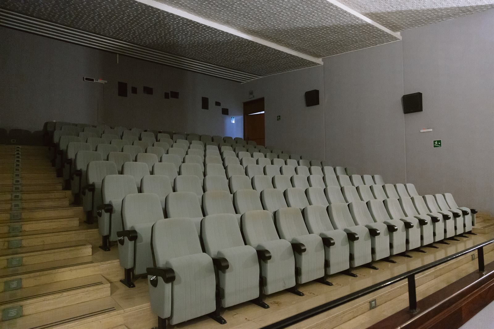 Platea a gradoni con poltrone chiare su pedane di legno, pareti grigie e soffitto forato